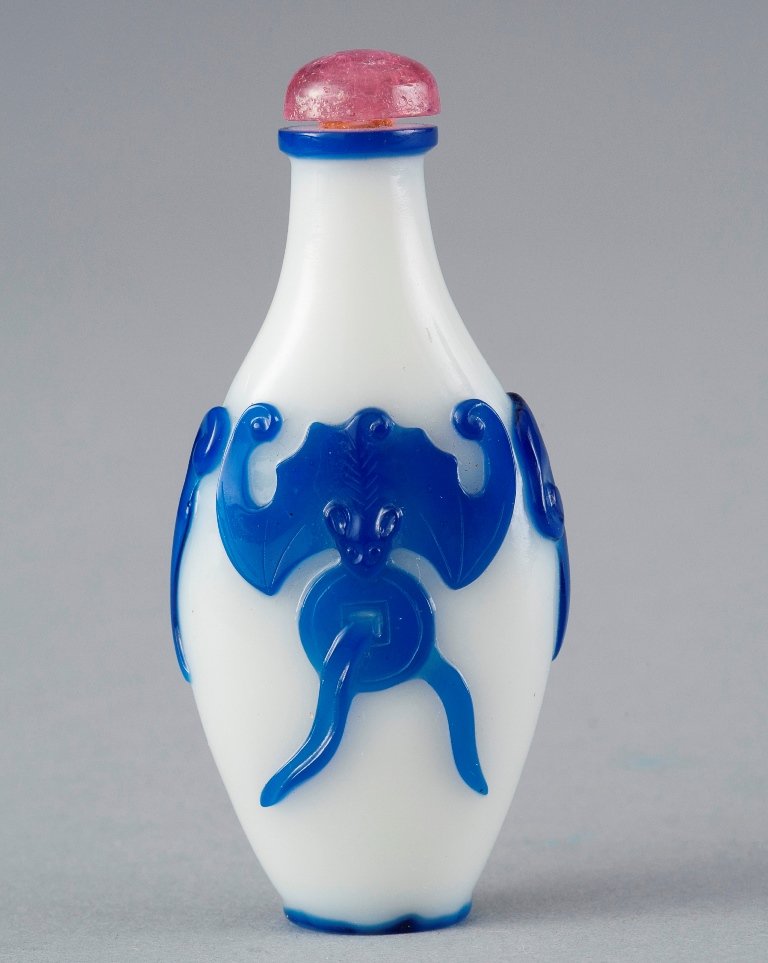 A BLUE OVERLAY WHITE GLASS SNUFF BOTTLE: 19TH CENTURY The bottle is pale white- even tone overall, the exterior is decorated with numerous sets of blue bats holding a spiritial coin. The cover of the bottle is pink colour tone. The edge of t