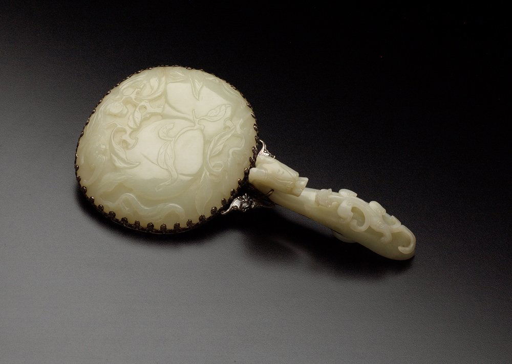 A WHITE JADE MOUNTED HAND MIRROR: 18TH CENTURY The handle is a pale greenish-white jade garment hook carved and undercut on top with a chilong crawling towards the dragon-head hook, and is attached to a silver mount on the underside.