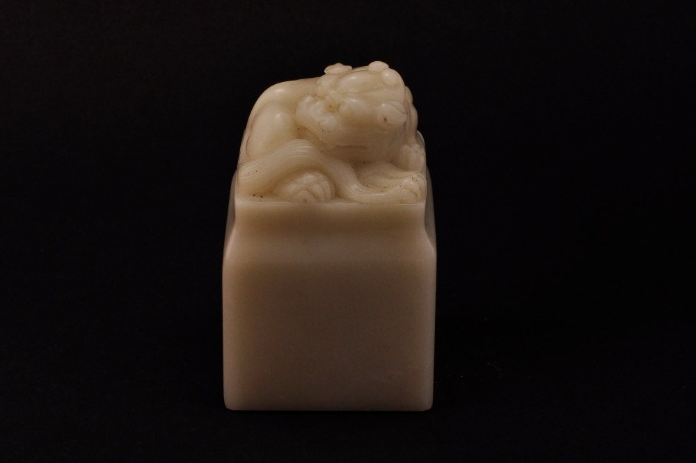 A SOAPSTONE CARVING OF LION SEAL: 民國 壽山石紙鎮 REPUBLIC PERIOD The soapstone is of white jade colour tone overall, the stone has a carving of a seated lion on the top. 2 3/8 in. (6 cm) tall