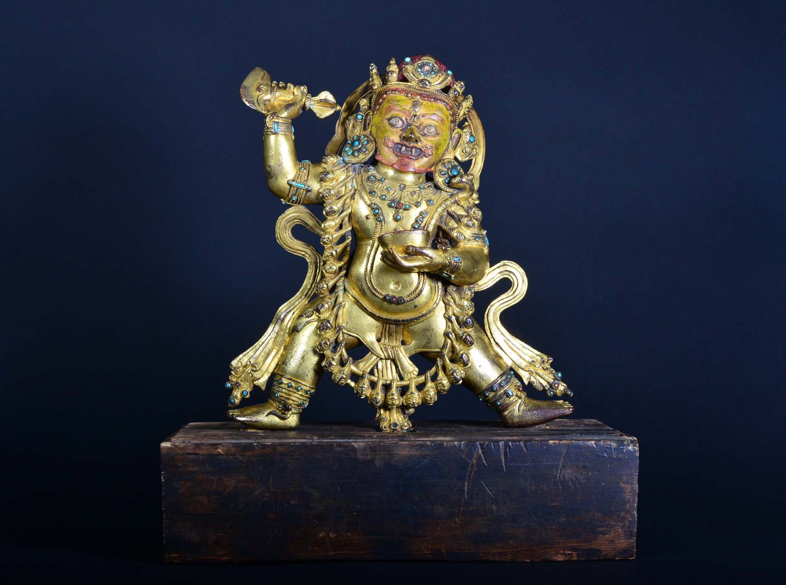 A Tibetan Gilt-Bronze Figure of Mahakala 15th - 16th Century: The figure is holding a chopper and skull cup in the primary hands, wearing a bone girdle, jewelry, and garland of severed heads, the ferocious face with bared fangs and three eyes, beneath the tall c
