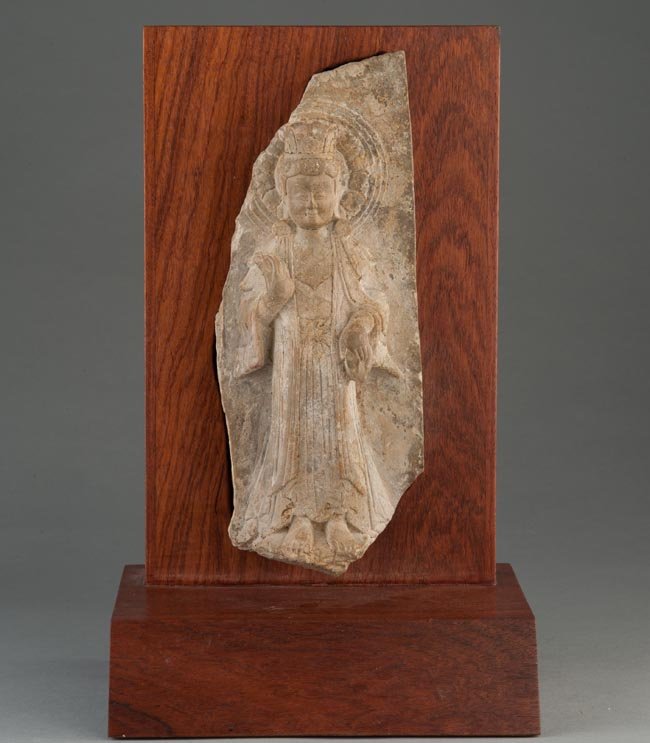 A STONE CARVING OF BUDDHA: NORTHERN WEI DYNASTY The stone carving is of a standing buddha wearing a long robe with the right hand in prayer postion. The stone carving is supported by a rectangular wooden stand. 10 1/4 in. (26 c