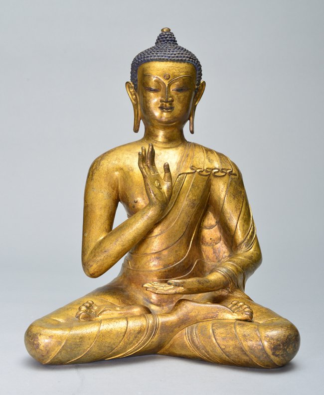 A LARGE GILT-BRONZE FIGURE OF BUDDHA: 16th Century Very finely cast seated in dhyanasana position. The Buddha's hands in the gestures of dhyana mudra and bhumisparsha mudra, wearing a long flowing robe over his left shoulder cascading dow