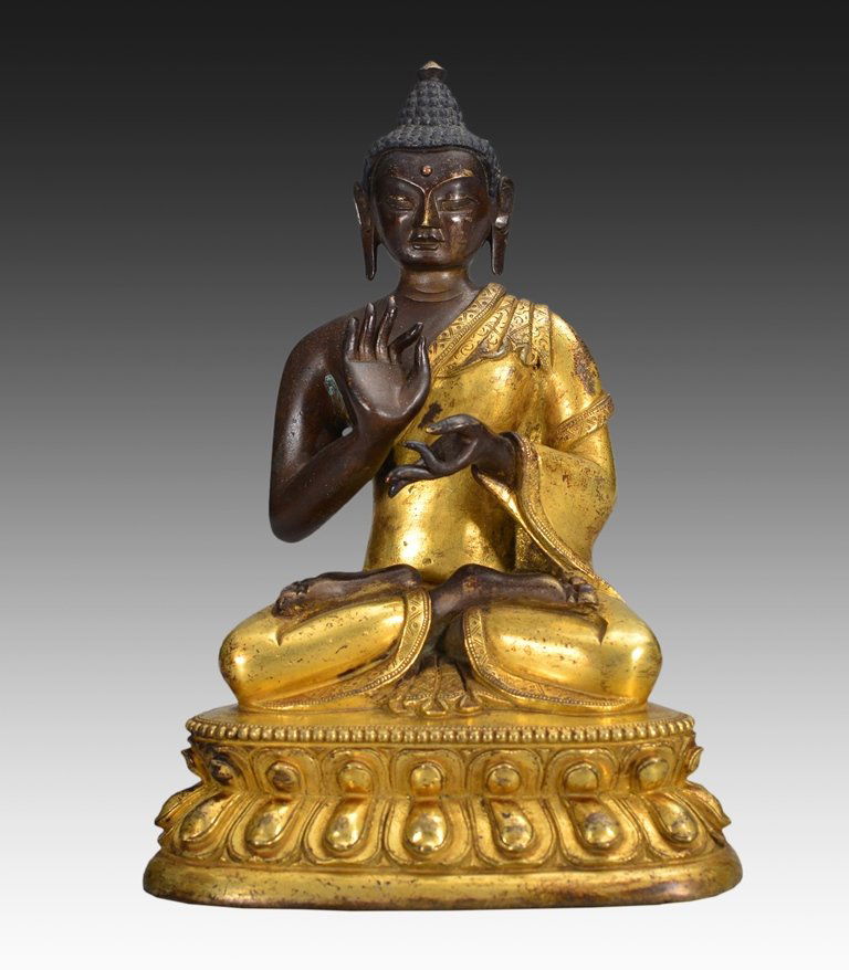 A BRONZE FIGURE OF BUDDHA: Seated on a double-lotus base, dressed in billowing patchwork robes that fall over the edge of the base, the face with bow-shaped mouth and elongated eyes flanked by pendulous earlobes, the hair in ti