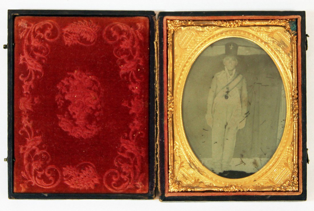 QUARTER PLATE AMBROTYPE OF MAN IN UNIFORM: In embossed leather case, photo shows an image of a young man wearing what is likely a Fraternal uniform, hand tinted cheeks, some scratching to surface. Size: 4-5/8 inches by 3-3/4 inches overall