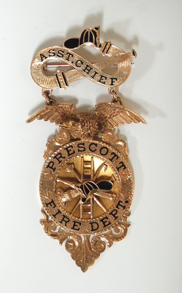 Prescott Fire Dept. Gold Suspension Badge: A beautiful highly detailed badge with hose and helmet ribbon over an eagle topped exhibition quality badge. Raised fire tool and ladder center in concave circle. Badge is 2 3/4” overall in height w