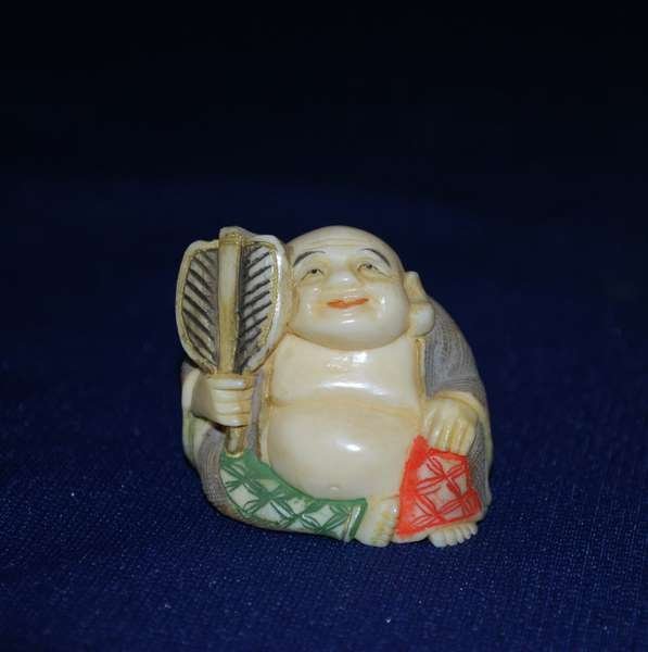 HAND CARVED IVORY NETSUKE CHARM: DEPICTING A BUDDAH FIGURED MAN SITTING WTH A FAN, INTRICATE CARVING, GREAT DETAIL COLOR, LOOP HOLES IN BACK, ORIENTAL SIGNATURE UNDER FOOT, SITS "1.25" HIGH