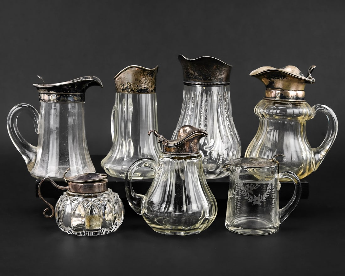 7-Piece Cut Glass Syrup Pitcher Lot with Stamped Lids: Group of seven cut glass syrup pitchers, each with a stamped metal lid. One is marked Gorham, another Hawkes. Most feature paneled sides, with one pitcher displaying an etched floral design.