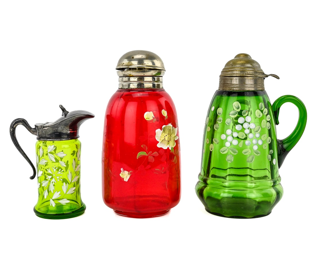 3 Enameled Ruby & Emerald Glass Pitchers: Three pieces of hand-painted enameled glass in ruby red and emerald green, each featuring floral decoration. The two taller pitchers have metal lids—likely zinc or tin—and measure approximately 7"