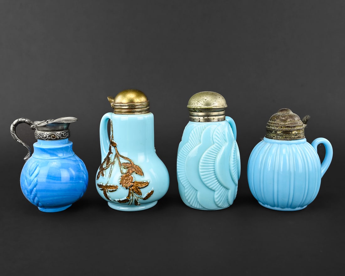 4 Pieces of Blue Milk Glass Syrup Pitchers: Group of four blue milk glass syrup pitchers, each with distinct decorative motifs. One features bronze-colored floral embellishments on a raised surface, while others are molded with designs such as 