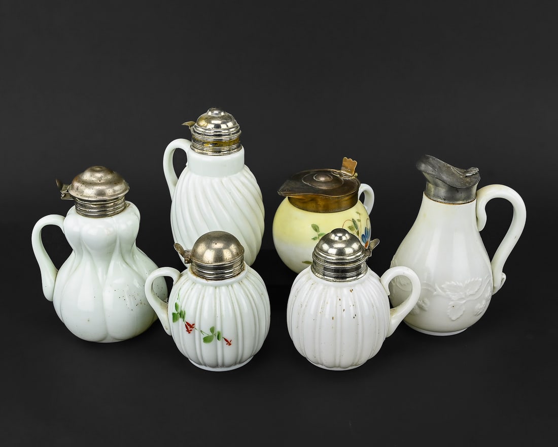 Six Pieces of Decorative Milk Glass Pitchers and Creamers: A collection of six milk glass pitchers and creamers featuring a variety of patterns including leaf and fern motifs, ribbed, and swirl designs. One piece is adorned with hand-painted floral decoration