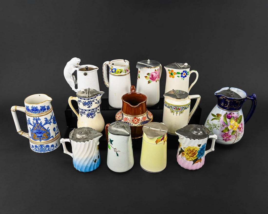 13 Porcelain Syrup Pitchers, Various Makers: Group of 13 porcelain syrup pitchers including a blue-handled Carlton Ware with pewter mid-hinged lid (approx. 6" tall x 4.5" wide); a Crown Staffordshire cream or yellow pitcher with small floral dec