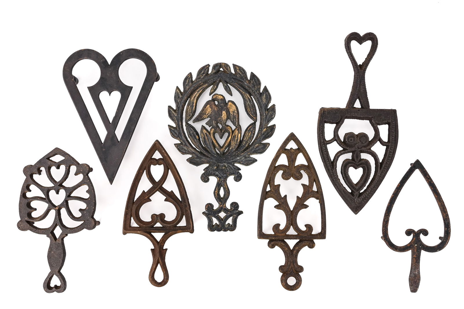 Group of 7 Cast Iron Decorative Trivets: Seven cast iron trivets featuring designs such as an eagle with heart in a wreath, hearts in a circular pattern, large spade-style trivets with hearts, stippled heart-decorated spade, and Gothic trace