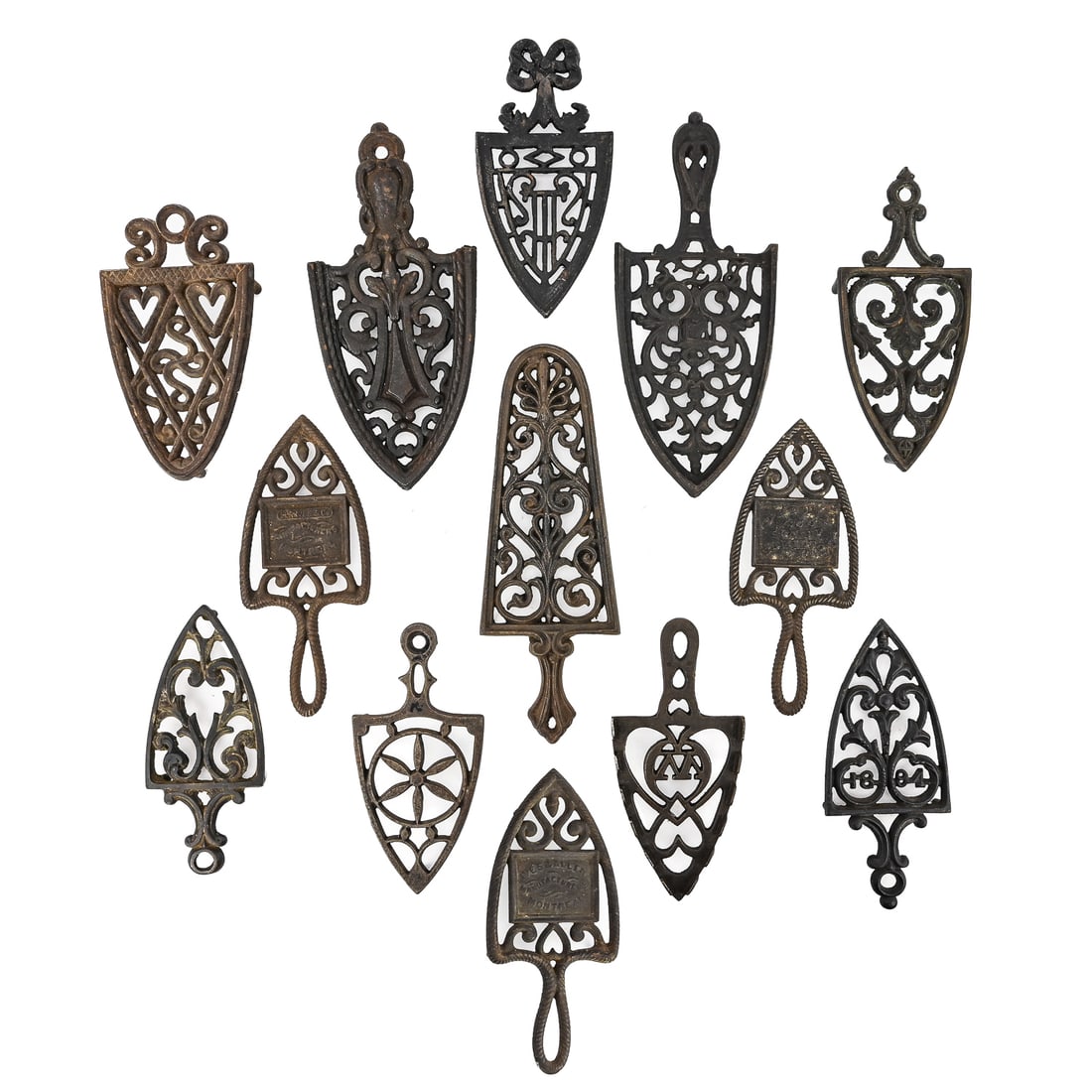 12 Cast Iron 20th-Century Decorative Trivets: Group of 12 cast iron trivets dating from the 20th century, featuring a variety of decorative motifs including fern and architectural patterns, harps, floral designs, and sigils. Several are marked wi