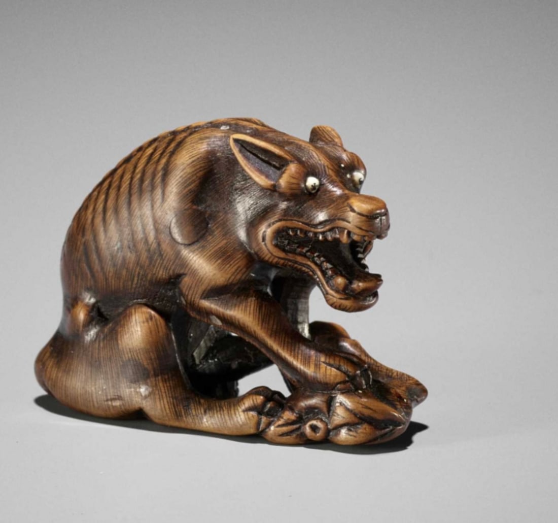 Fine Japanese Netsuke by Masatomo: Carved as an emaciated, snarling wolf with neatly incised spine and fur, large, pricked ears, and eyes inlaid in bone with black pupils, the tail tucked underneath, gaurding a haunch of venison under