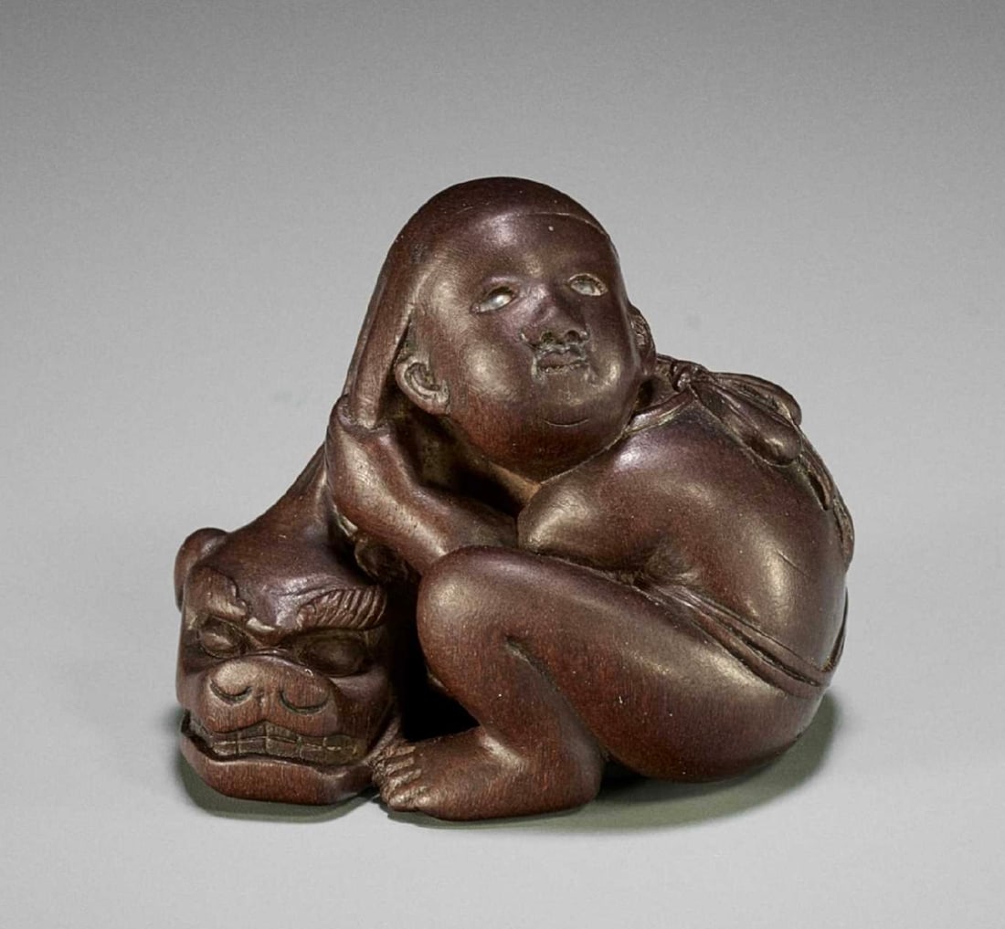 Fine Netsuke Signed with a Kakihan: Well carved as a seated boy removing his shishimai mask, the flexible fabric of the mask superbly executes as the boy pulls the mask off his head, his face with a fine expression and glass-inlaid eyes