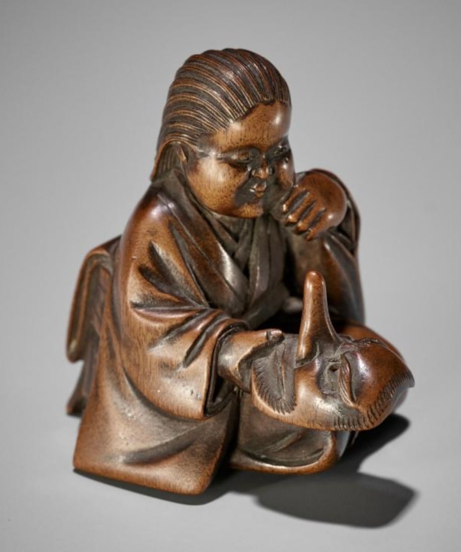 A Fine Wood Shunga Netsuke of Okame Holding a Konoha Tengu Mask: Okame dressed in a heian period robe, kneeling and smilins while holding in her right hand a konoha-tengu mask, its long nose pointing up. Her hair tied back into a loose knot, and the pupils are inla