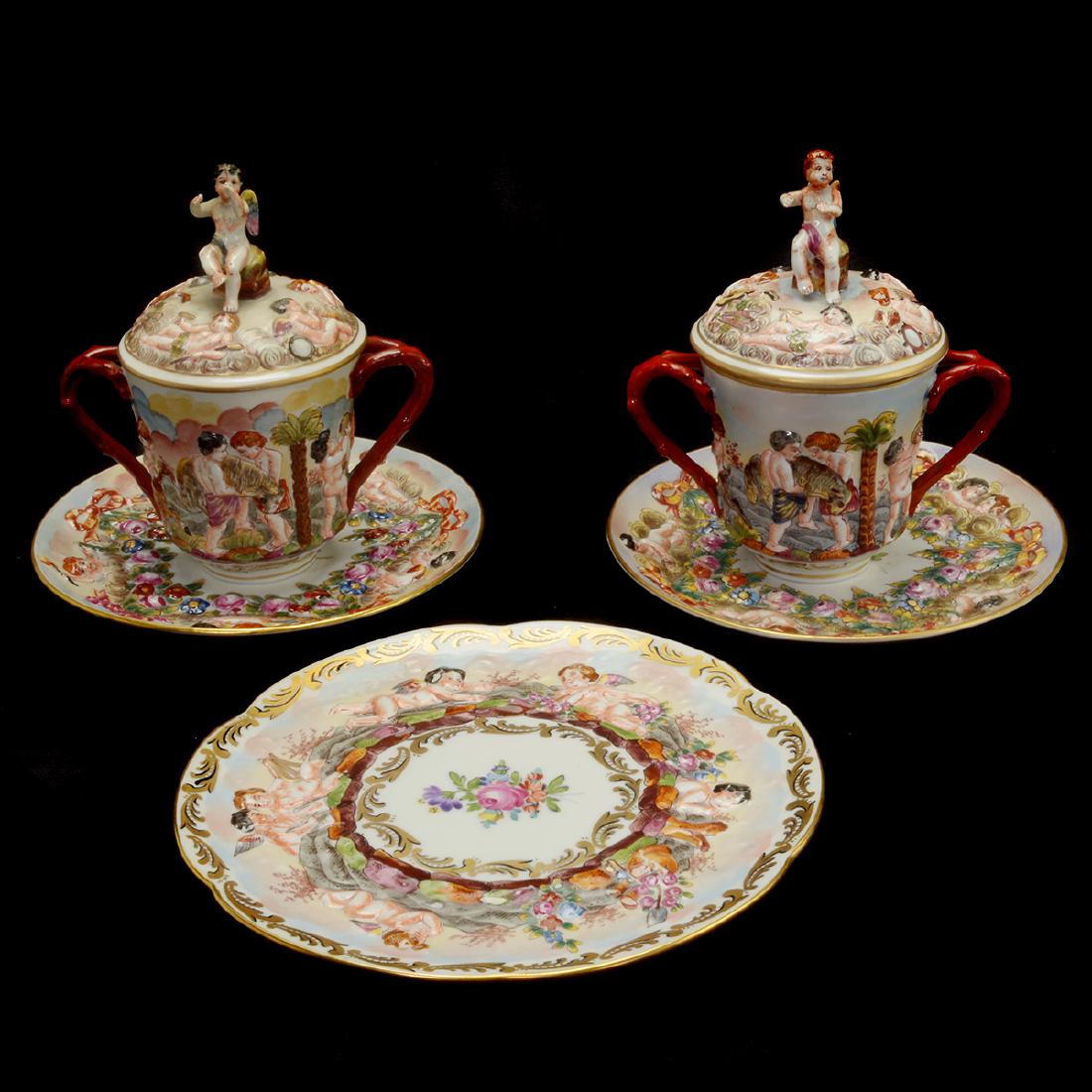 Pair of Capodimonte Porcelain Lidded Cups with Saucers,: Pair of Capodimonte Porcelain Lidded Cups with Saucers, Additional Dish. Each lidded cup with a putto form finial, two twig form handles, and decorated in relief with cherubs and satyrs performing var