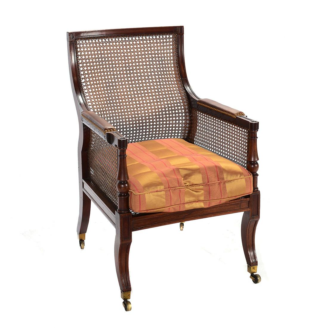 William IV Mahogany and Cane Library Chair.: William IV Mahogany and Cane Library Chair. Curved cane back; the frame with carved coffering; riveted leather padding to the arms; baluster carved arms supports; on casters; red and gold silk upholst