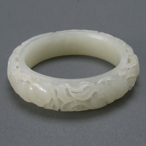 A Carved White Jade Bangle: A Carved White Jade Bangle A solid interior band supports intricately carved openwork design of scrolling vines, leaves and a peach, entwining bats throughout. Diameter 3 inches