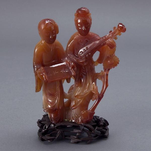 A Carnelian Carving of Two Female Figures: A Carnelian Carving of Two Female Figures, each holding a lute and scroll, attached to a reticulated wood stand {Height 5 3/4 inches}