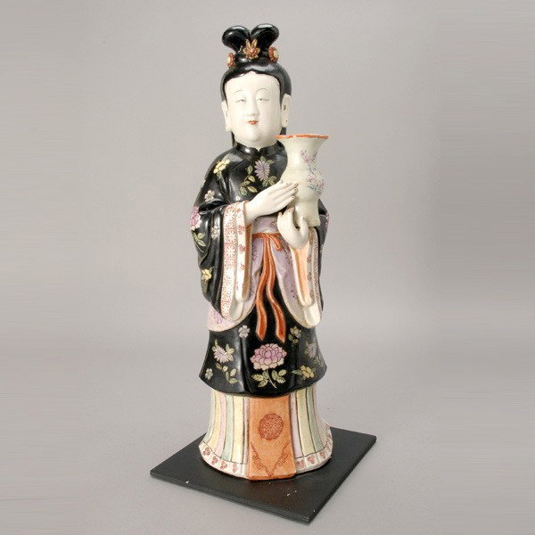 A Polychrome-Enameled Porcelain Figure: A Polychrome-Enameled Porcelain Figure, Late Qing/Republic Period, shown wearing black-ground floral robes while holding a vase, fixed to a square base {Height 19 inches}