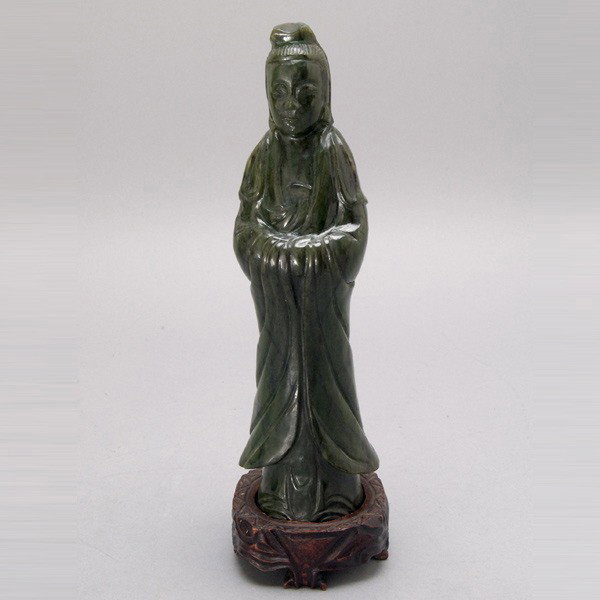 A 'Spinach' Jade Guanyin: A 'Spinach' Jade Guanyin, shown standing upon a matching wood stand, the long robes displaying dark black inclusions {Height 9 3/4 inches} [Pinpoint chips to edges and shoulder of robe]