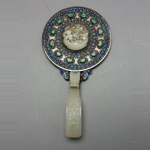 A Jade Mounted Hand Mirror, Qing Dynasty: A Jade Mounted Hand Mirror, The circular mirror back set with a central carved jade plaque depicting a figure standing on a large fish, surrounded by concentric patterns of finely enameled floral mean