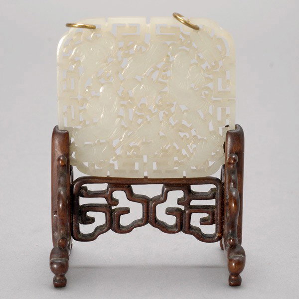 A Finely Carved Jade Plaque: A Finely Carved Jade Plaque, the reticulated square shaped panel depicting two figures amidst lotus flowers, birds and bats, each top corner with a gilt metal ring; displayed upon a matching carved wo