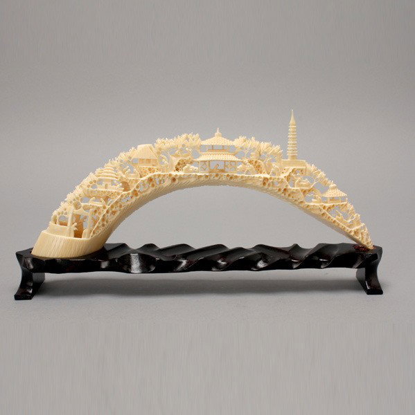A Carved Ivory Tusk*: A Carved Ivory Tusk*, the arched form intricately carved with a figural landscape amidst trees and buildings, with a matching wood stand {Length: 12 1/4 inches (31 .1 cm)}