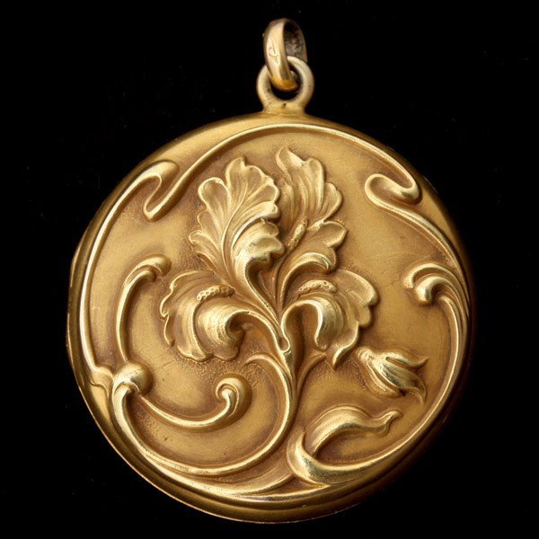 10K YELLOW GOLD ART NOUVEAU LOCKET.: 10K YELLOW GOLD ART NOUVEAU LOCKET. Featuring a repousé iris design with monogram back, measuring approximately 32 mm in diameter, completed by a gold bail. 5.40 dwts