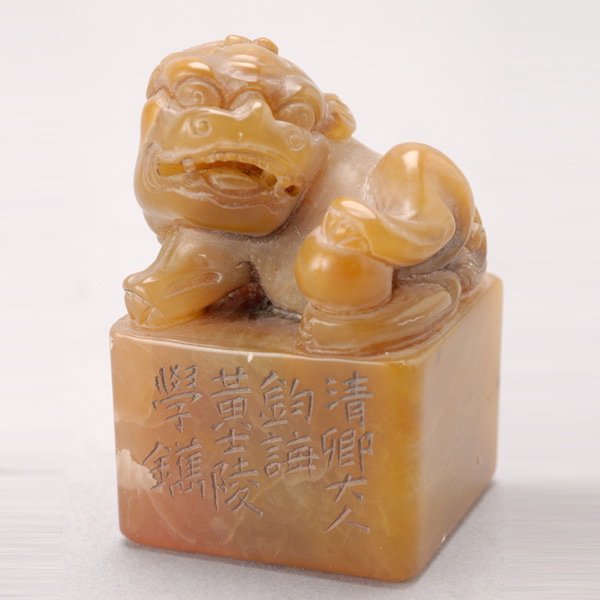 A Soapstone Seal: A Soapstone Seal, The orange stone of square form, surmounted with a playful lion holding a small ball between its hind legs, the seal face carved in yin form with four characters reading 'cong wu suo