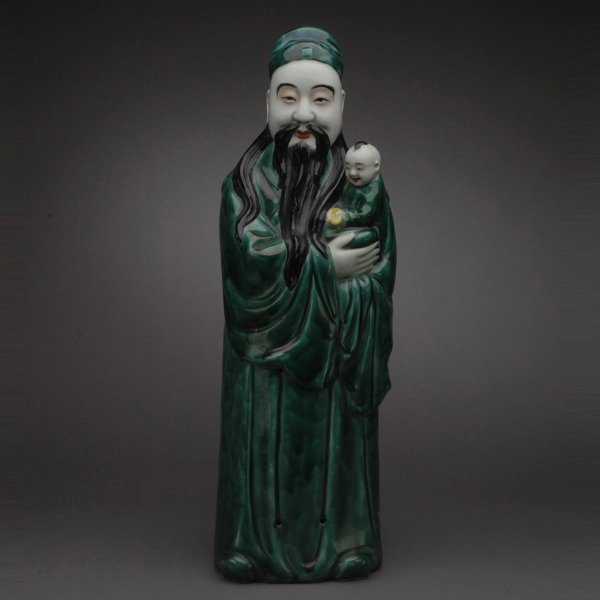 A Glazed Porcelain Star God: A Glazed Porcelain Star God, with a long black beard, standing while holding a child, wearing dark green robes with a matching hat {Height: 24 1/2 inches (62.2 cm)} [Chips]