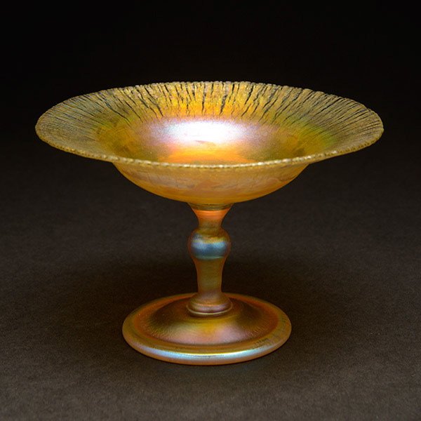 Tiffany Studios Favrile Glass Compote: Tiffany Studios Favrile Glass Compote. Executed in iridescent gold Favrile glass with purple and pink highlights. The upper section rising from a circular foot on a slender stem which is knobbed neari