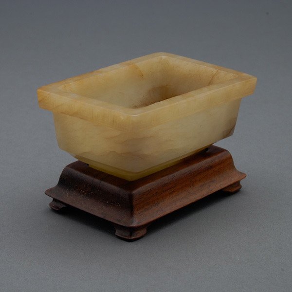 A Small Jade Incense Burner, Qing Dynasty: A Small Jade Incense Burner, The rectangular vessel with a deep well, the outer edges of the rim incised with a keyfret band, all on a tall recessed base, with a fitted wood stand. Qing Dynasty, Width