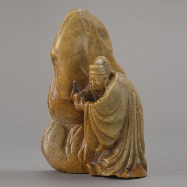 A Carved Soapstone Figural Boulder, Qing: A Carved Soapstone Figural Boulder, Qing Dynasty, carved in relief with a scholar holding a rectangular black tablet and dressed in long flowing robes, shown standing in front of a large rock {Height: