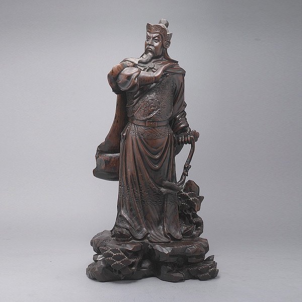 A Wood Carving of Guandi: A Wood Carving of Guandi, the bearded figure shown standing upon a base of rocks and pine branches, wearing a dragon robe and with one hand holding his sword {Height: 21 1/2 inches (55 cm)} [Chips, fi