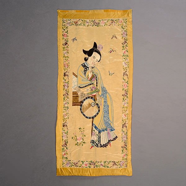An Embroidered Wall Hanging of a Beauty, Late Qing: An Embroidered Silk Wall Hanging of a Beauty, The coy female figure shown holding a palm leaf fan and calligraphy while wearing long floral robes against a pale yellow ground, her hair pinned back in