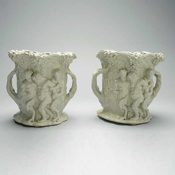 Pair Staffordshire Ironstone Urns: Pair of Staffordshire Bacchanalian Ironstone Urns, each cylindrical form with faux bark indentations and tree branch handles, continuously decorated with Bacchus being supported by a couple of satyrs 