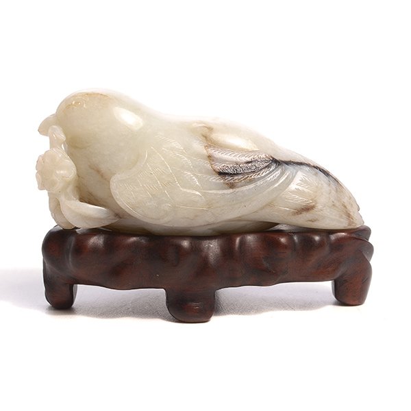 A Jade Bird: A Jade Bird Carved as a resting quail grasping a floral branch, the white colored jade with black and russet inclusions, with a fitted wood stand. {Length: 3 1/8 inches (7.9 cm)}