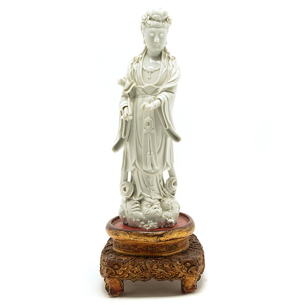 A Dehua Figure of Guanyin, Late 19th/Early 20th Century: A Dehua Figure of Guanyin Modeled standing on a dragon in stylized waves, the arms lowered supporting a lotus stem, dressed in loose fitting robes falling into elegant folds and opening at the chest r