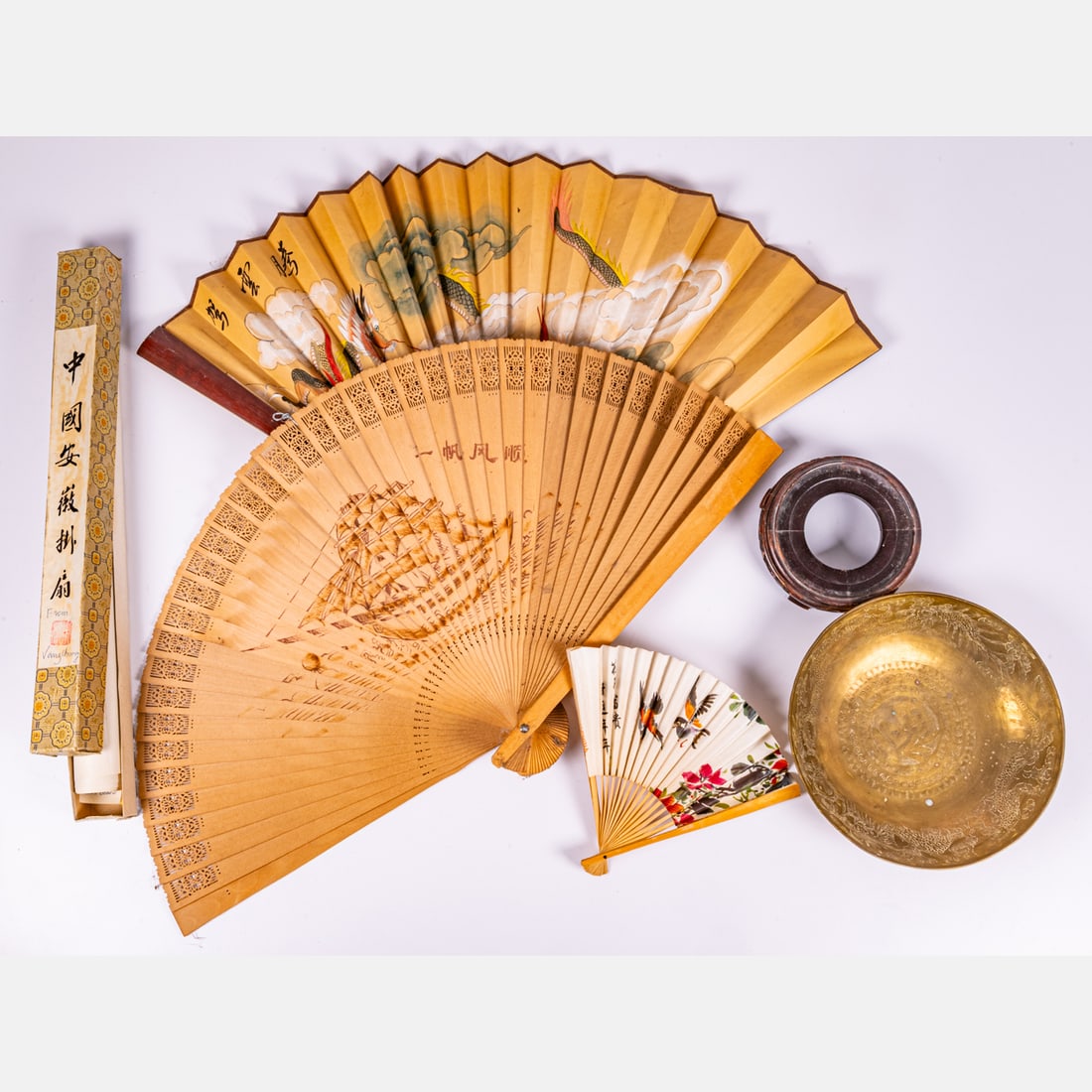 A Group of Asian Decorative Objects: Three Asian Fans, Depicting Dragons and Birds, One fan is bamboo depicting a tall masted ship.Together with an etched brass bowl depicting dragons with a stand and a painted scroll in original box. La