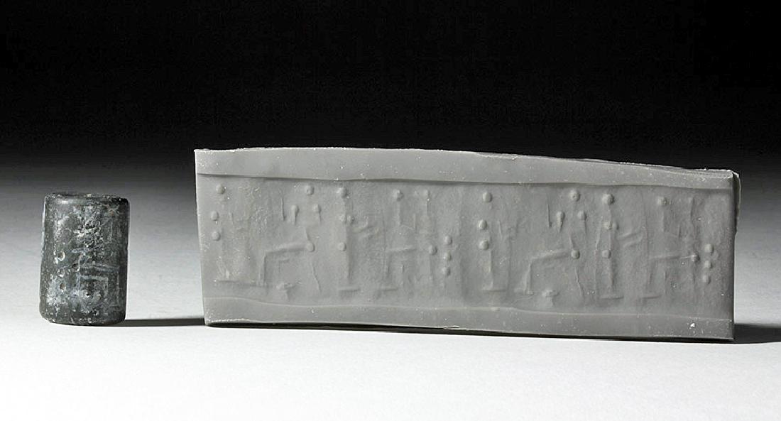 Mesopotamian Cylinder Seal, ex-Christie's, Royal Athena: Ancient Near East, modern day Syria/Turkey, Kingdom of Mitanni, ca. 1350 to 1300 CE. A smoothed black stone cylinder seal, pierced through for suspension, that shows two standing worshippers before tw