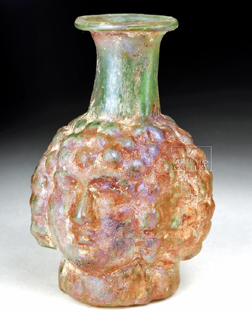 Roman Glass Head Flask - Possibly Dionysos as a Boy: Roman, Imperial Period, ca. late 2nd century CE. A striking mold-blown head flask comprised of translucent green glass with an aubergine tinge on the face and coiffure. The vessel presents a youthful 