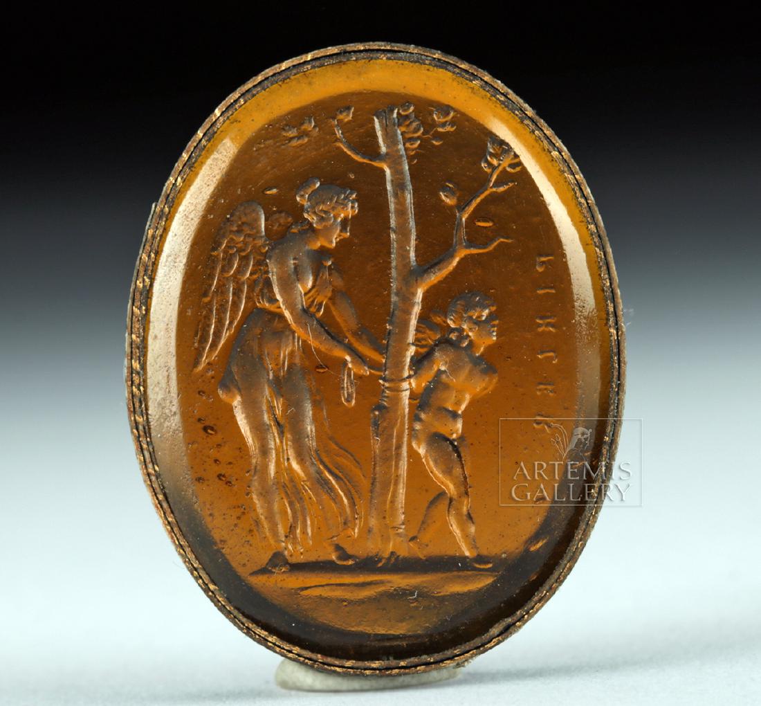Neoclassical Glass Intaglio - Nike & Eros: Northern Europe, Neoclassical period, ca. 18th to 19th century CE. An attractive amber-hued glass intaglio depicting a winged Nike tying Eros to or untying him from a tree, with a Greek inscription be