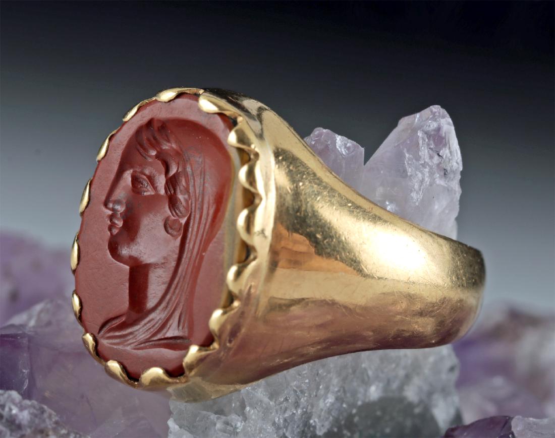 18K Gold Ring w/ Roman Jasper Intaglio of Woman: Roman, Imperial Period, Julio Claudian, ca. 1st century CE. A lovely ovoid intaglio depicting the left-facing head of a woman, hand-carved from crimson-hued jasper. Her visage is composed of a singula