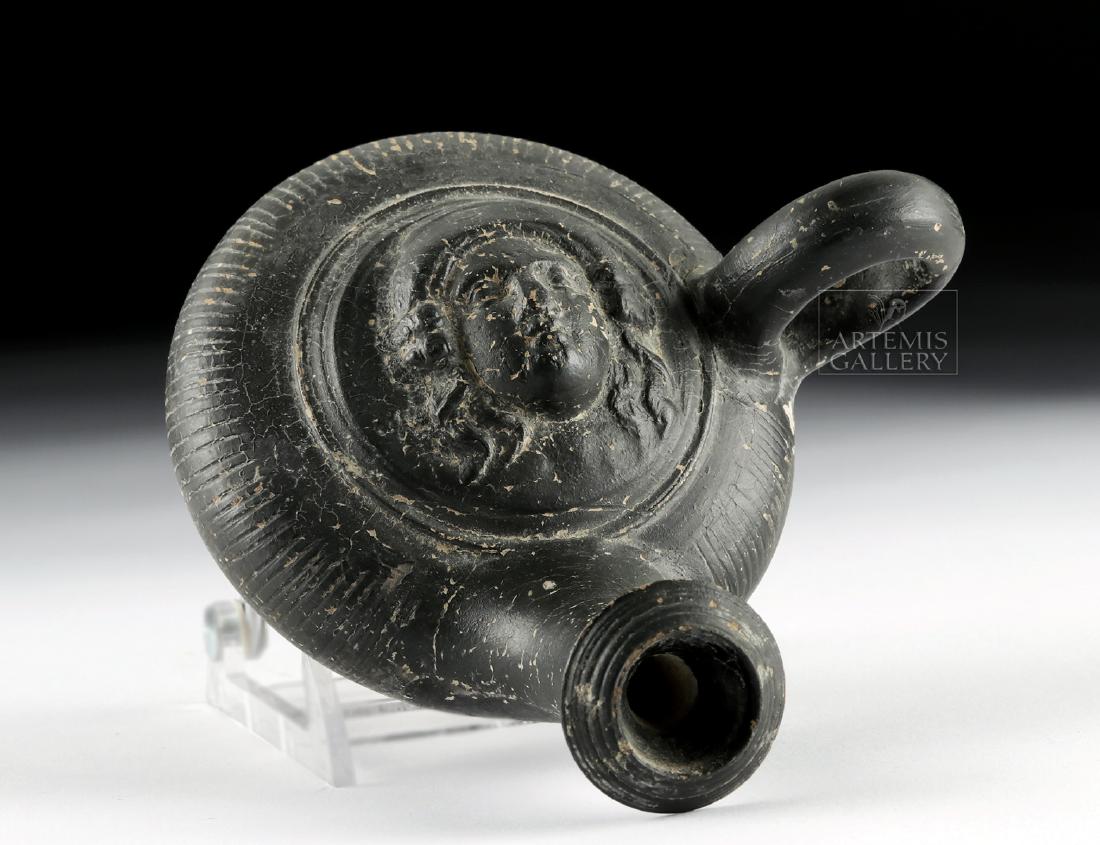 Greek Campanian Blackware Guttus w/ Female Face: Magna Graecia, Southern Italy, Campania, ca. 360 to 320 BCE. A lovely blackware guttus (or guttos) form with a rounded body, a single loop handle, and a projecting spout with a funnel mouth, all upon