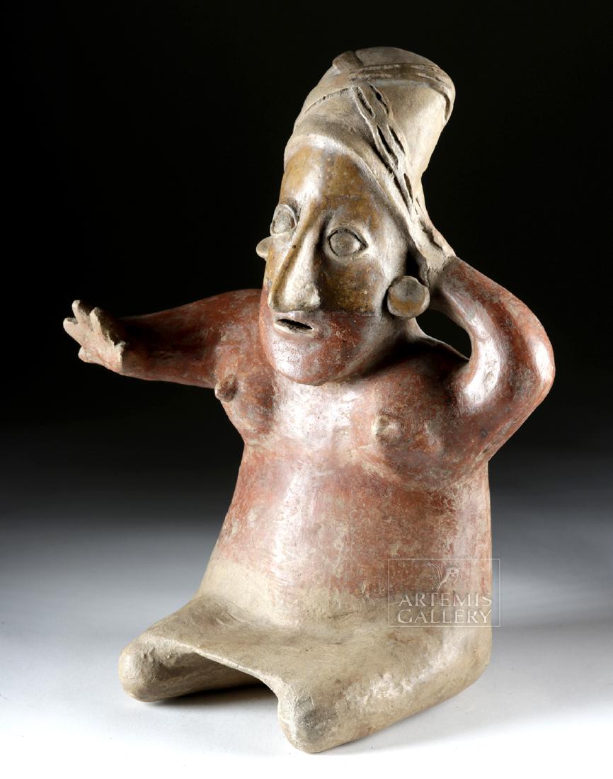 Jalisco Redware Seated Female Figure: Pre-Columbian, West Mexico, Jalisco, ca. 300 BCE to 300 CE. A hand-built pottery figure seated upon attenuated legs and wearing a knee-length skirt. Her nude upper body boasts ample breasts and rounde