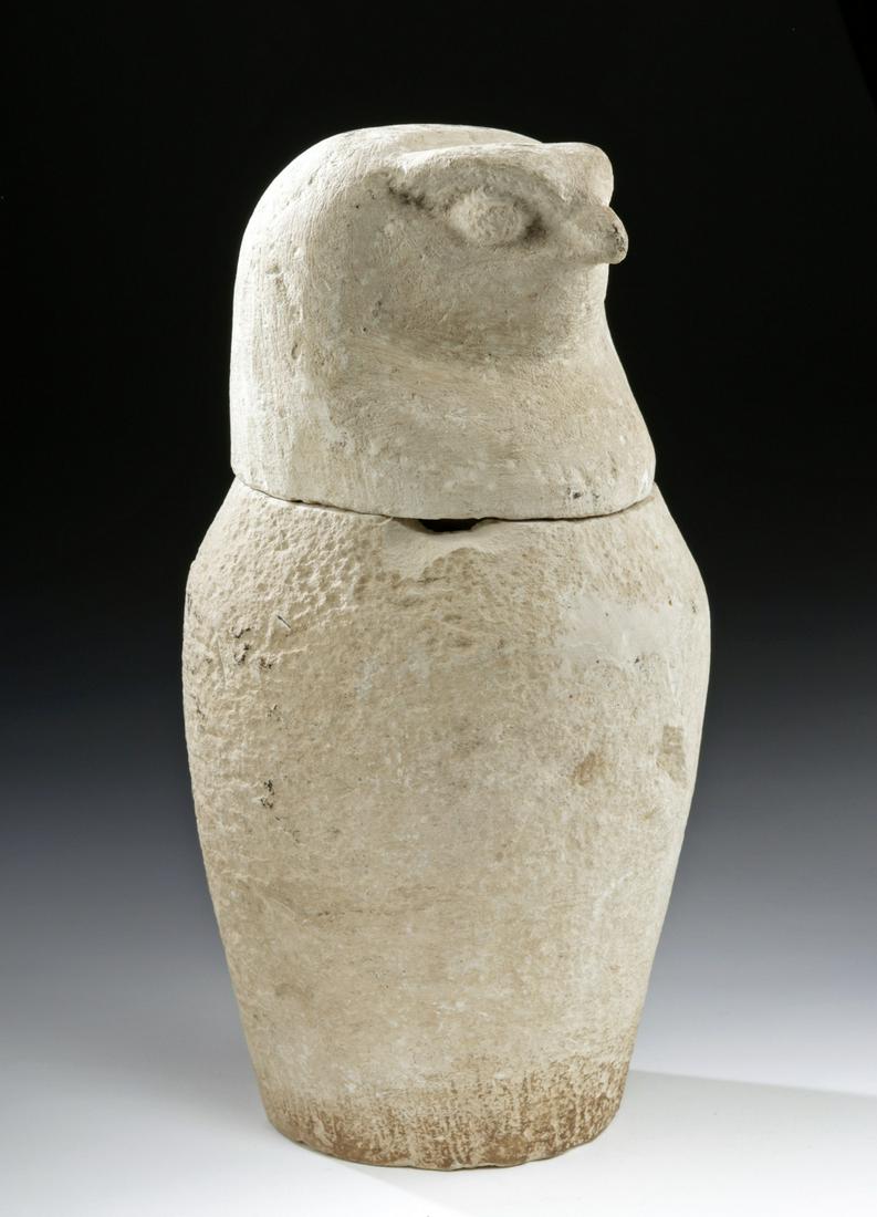 Egyptian Limestone Canopic Jar w/ Falcon Lid: Egypt, Late Dynastic Period, 26th to 31st Dynasty, ca. 664 to 332 BCE. A hand-carved limestone canopic jar of an elegant minimalist presentation. The cylindrical vessel has a flat base, an inverted pi