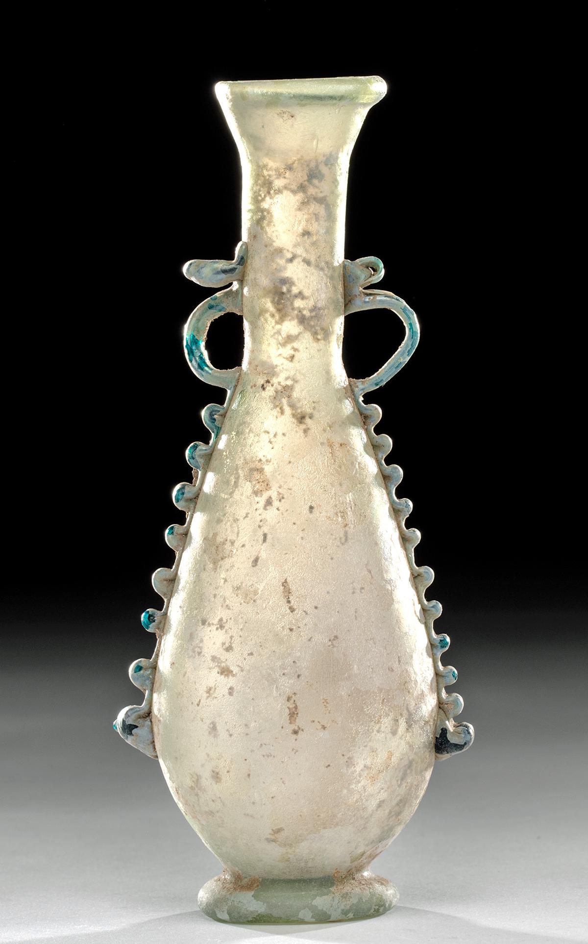 Roman Glass Amphoriskos w/ Trailing Handles: Roman, Imperial Period, ca. 1st to 3rd century CE. A delicate, lovely free blown amphoriskos, made of a very pale green, translucent glass, with thin silvery and pearlescent iridescence on the surface
