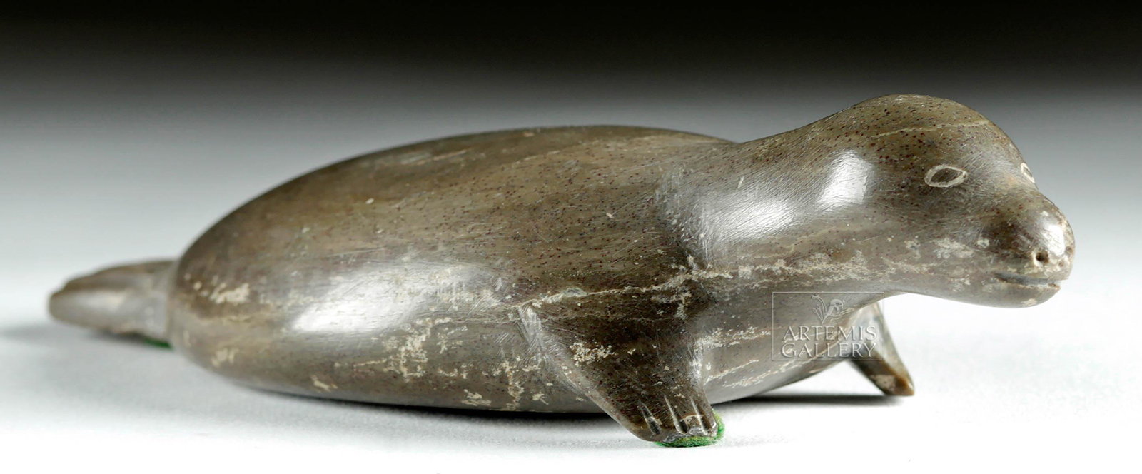 20th C. Canadian Inuit Soapstone Seal Carving - Okhena: Native American / First Nations, northern Canada, Inuit, ca. 1920s or later CE, signed by "Okhena". A handsome mottled grey and grey-green soapstone carving of a seal, its body elongated and widened i